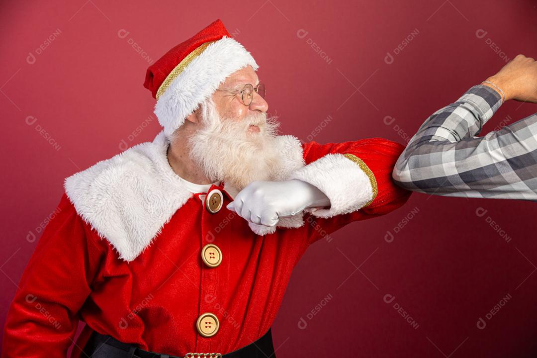 Verdadeiro Papai Noel com fundo vermelho, usando óculos e chapéu fazendo saudação de cotovelo. Natal com distanciamento social. Covid-19