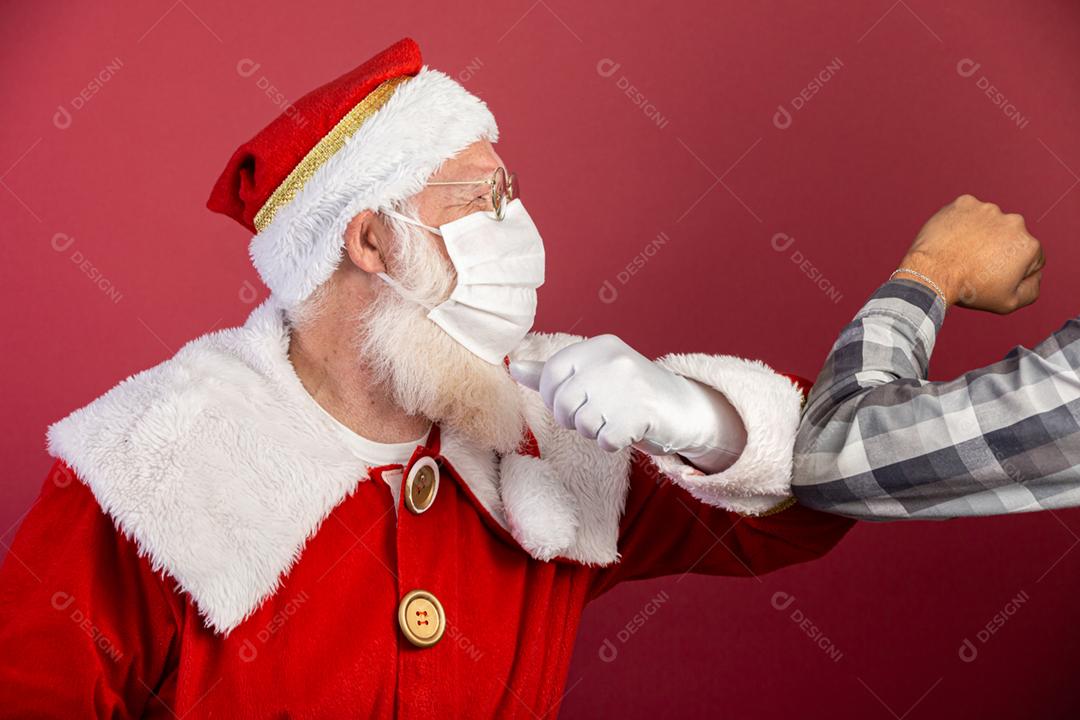 Verdadeiro Papai Noel com fundo vermelho, usando óculos e chapéu fazendo saudação de cotovelo. Natal com distanciamento social. Covid-19