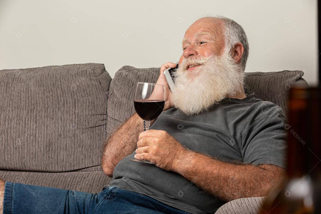 Homem barbudo elegante e bonito bebendo vinho tinto e ligando para o telefone em casa