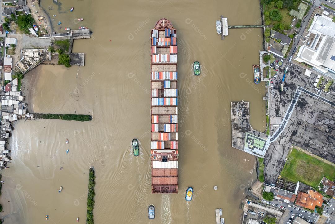 Vista aérea de um grande cargueiro de contêineres se dirige ao mar auxiliado por rebocadores