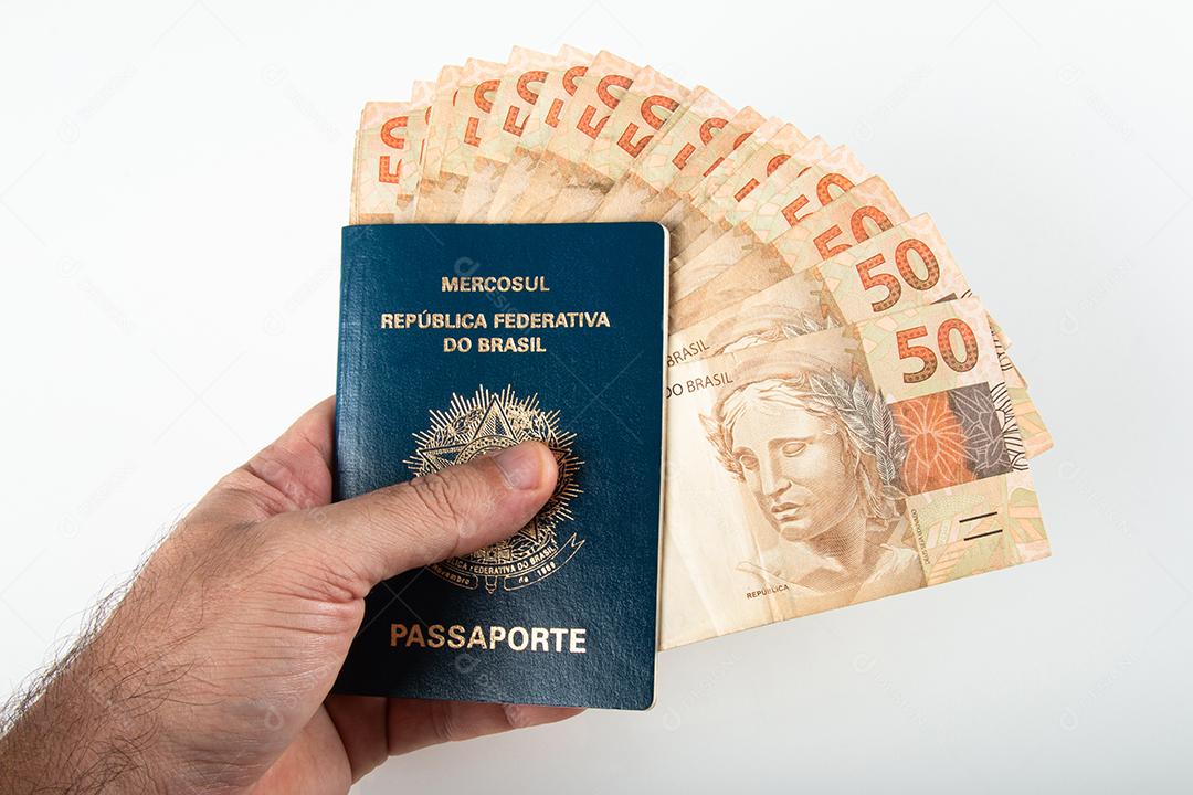 Mão segurando passaporte brasileiro com notas de cinquenta reais. Brasil
