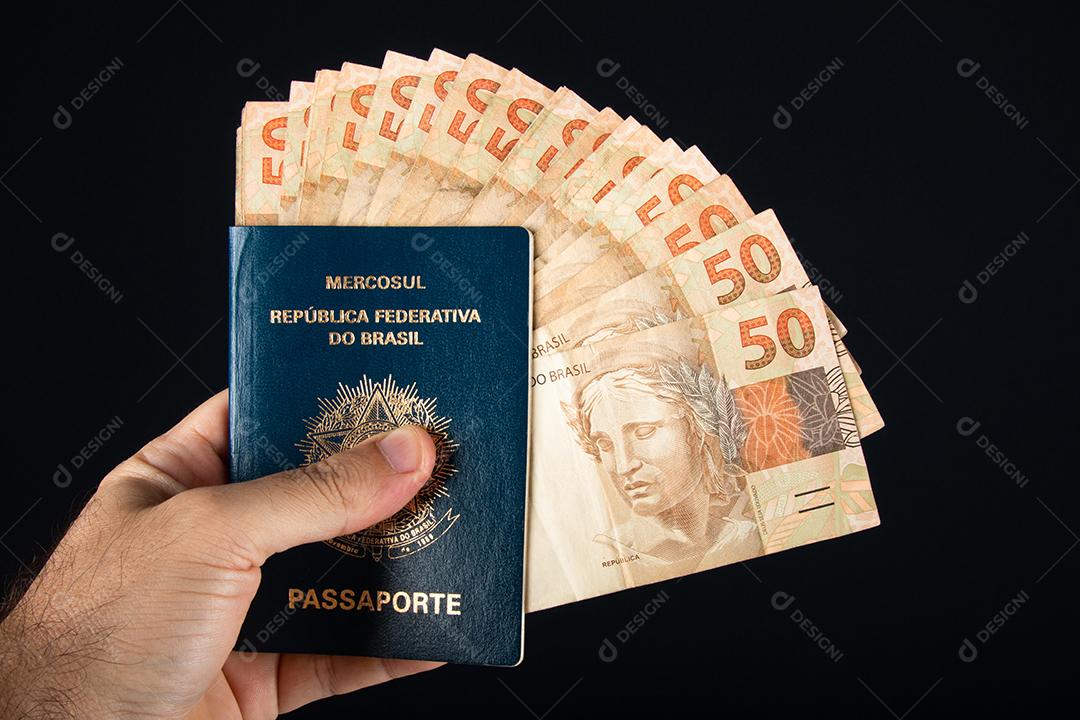 Mão segurando passaporte brasileiro com notas de cinquenta reais. Brasil