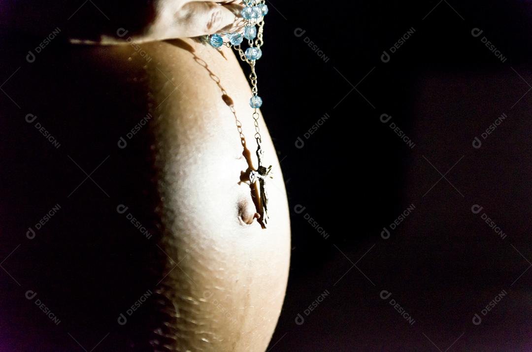 Grávida com as mãos na barriga e rosário, rezando.