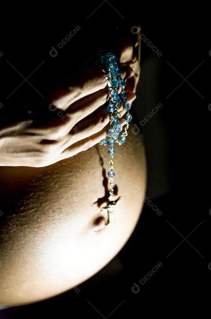 Grávida com as mãos na barriga e rosário, rezando.