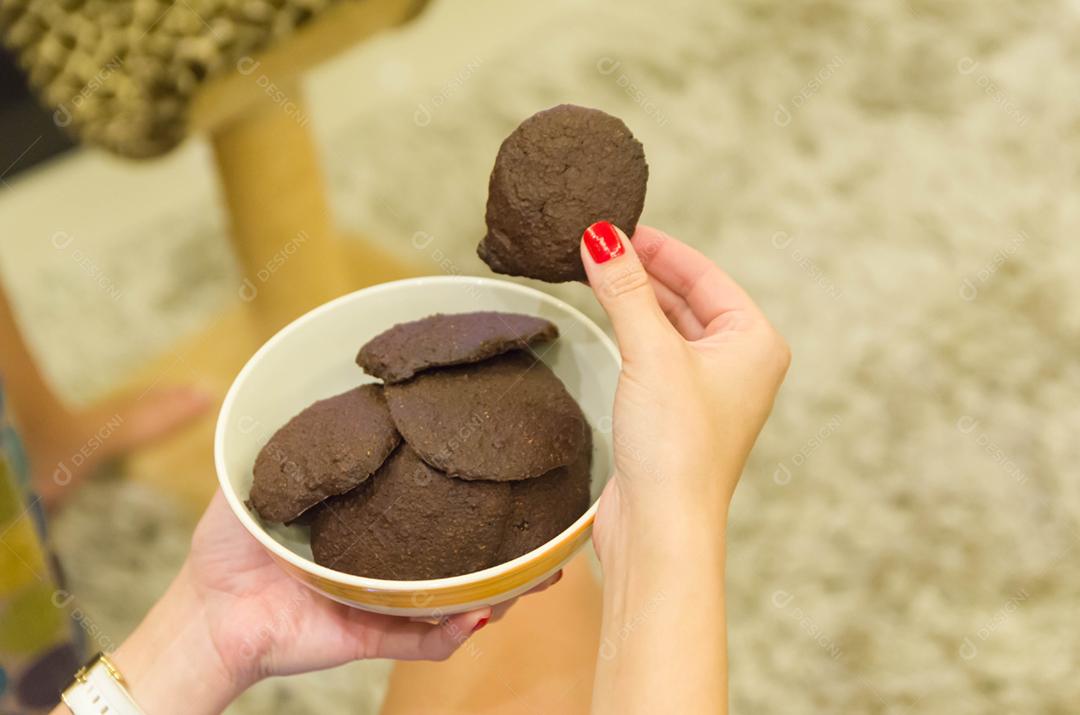 Mão de mulher segurando biscoito de chocolate caseiro em uma tigela