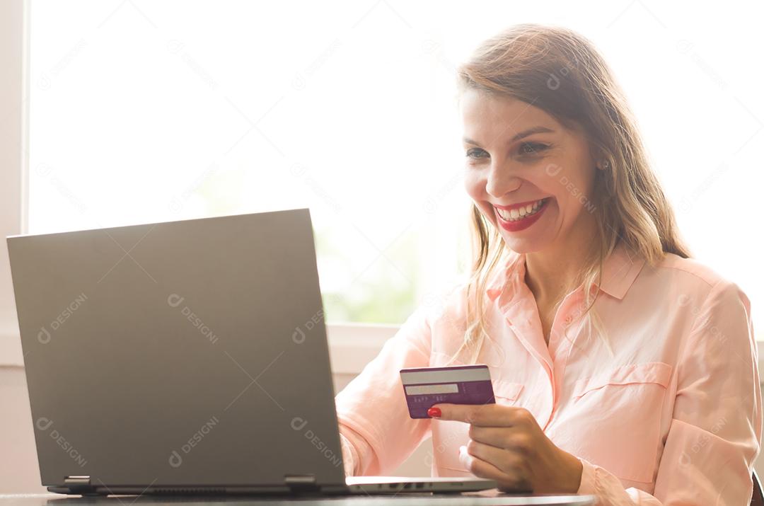 Jovem loira caucasiana feliz comprando na internet, cartão de crédito na mão.