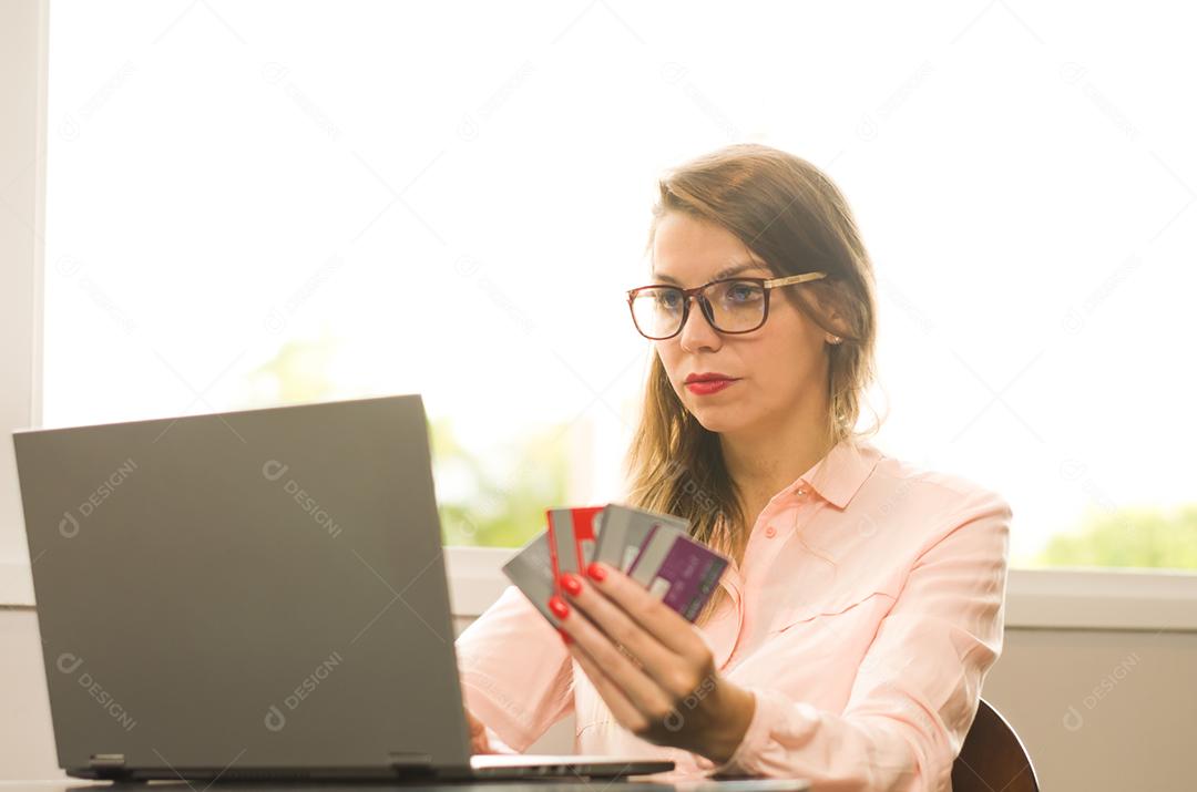 Jovem loira caucasiana preocupada em pagar contas eletrônicas, com vários cartões de crédito na mão.