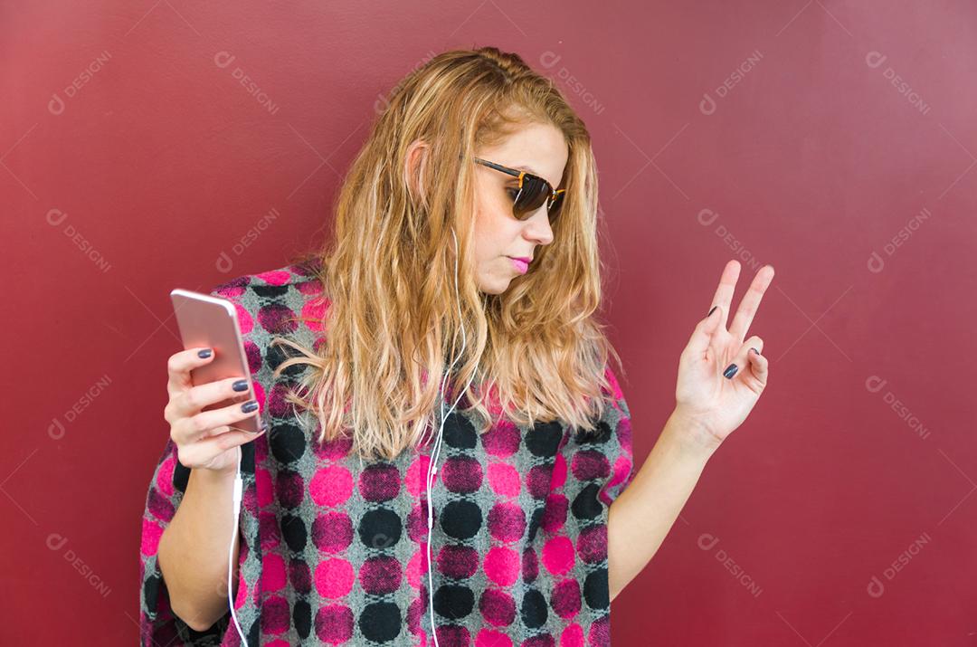 A jovem bela loira sorridente com fones de ouvido, ouvindo a música e dançando na frente do fundo vermelho.