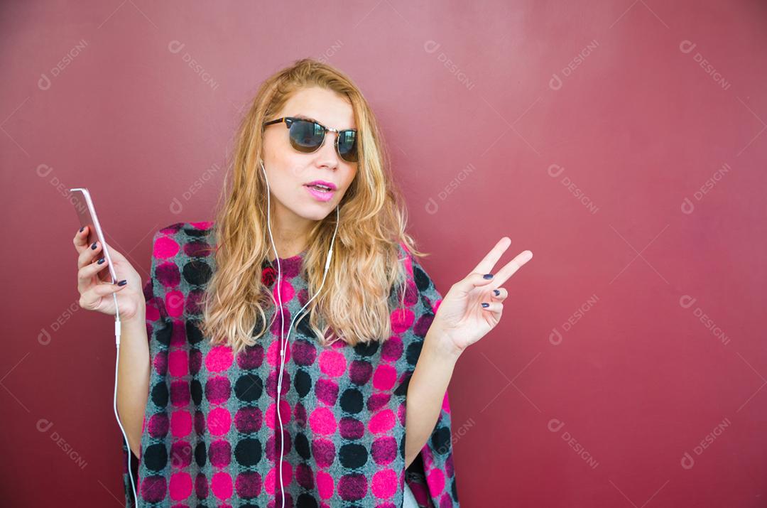 A jovem bela loira sorridente com fones de ouvido, ouvindo a música e dançando na frente do fundo vermelho.