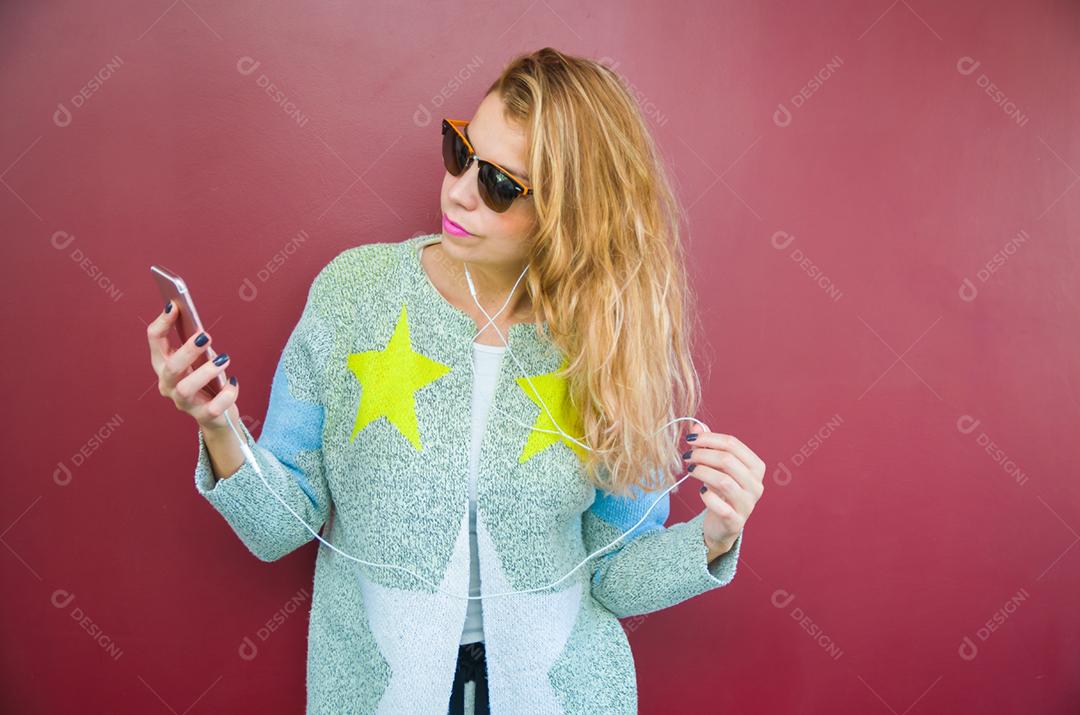A jovem bela loira sorridente com fones de ouvido, ouvindo a música e dançando na frente do fundo vermelho.