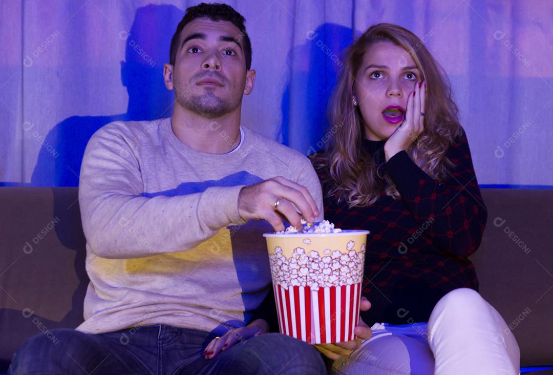 Jovem casal assistindo filme de suspense no quarto escuro com pipoca no sofá totalmente atenção