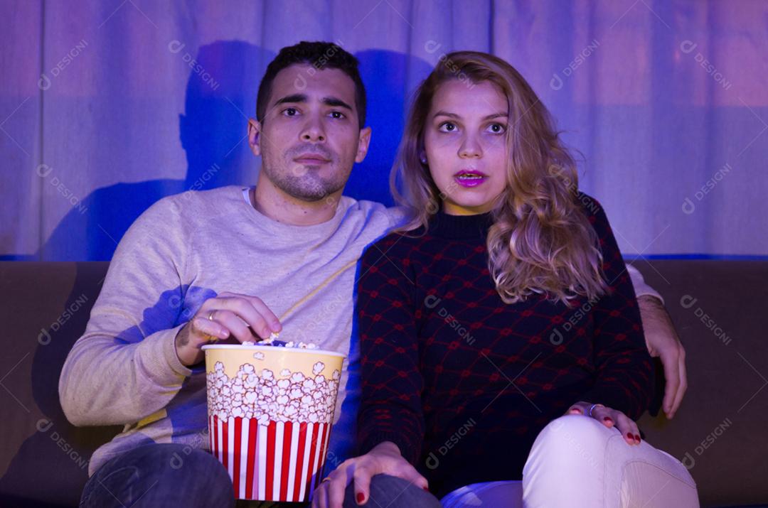 Jovem casal assistindo filme de suspense no quarto escuro com pipoca no sofá totalmente atenção