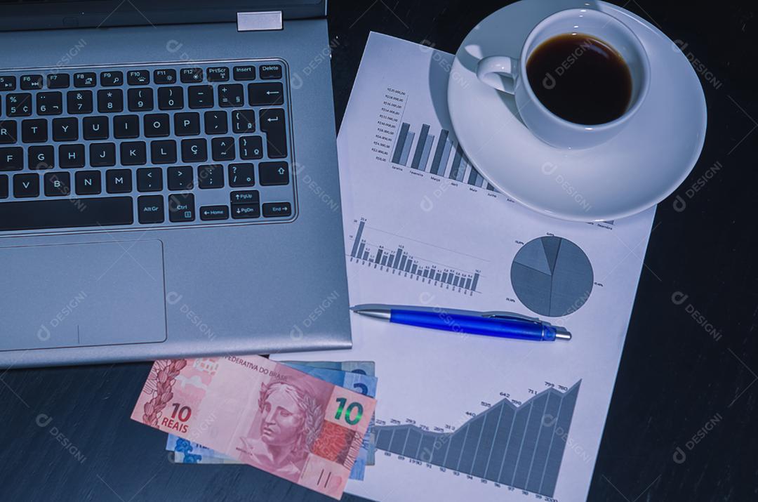 Computador portátil, xícara de café, gráficos de rendimento, caneta e moeda do Brasil.