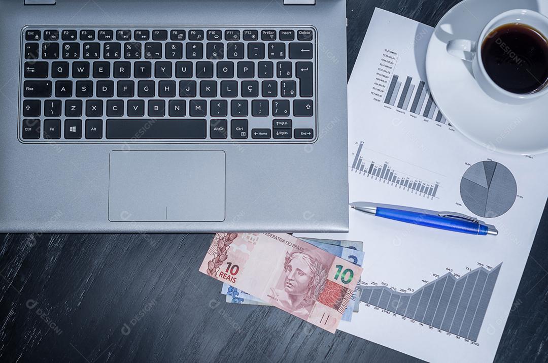 Computador portátil, xícara de café, gráficos de rendimento, caneta e moeda do Brasil.