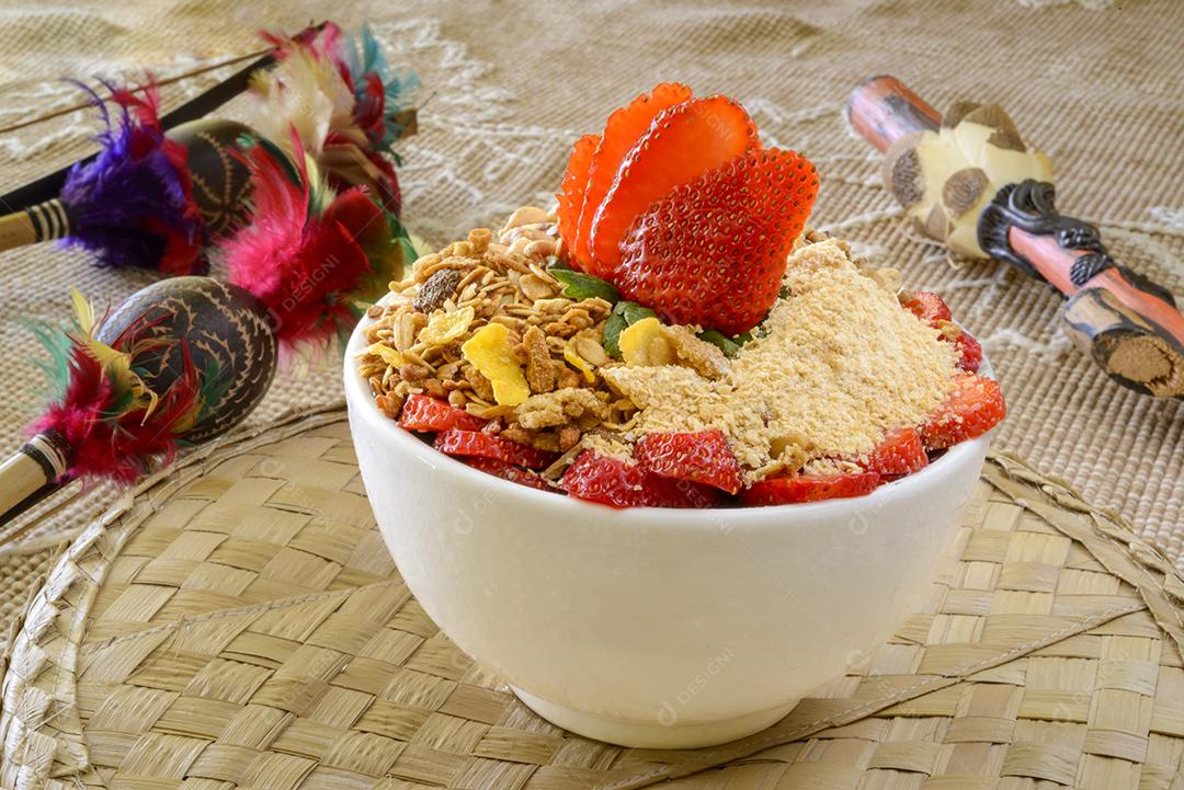 Tigela de açaí brasileiro com morangos, granola e amendoim em uma mesa com artesanato da região amazônica borrada ao fundo.