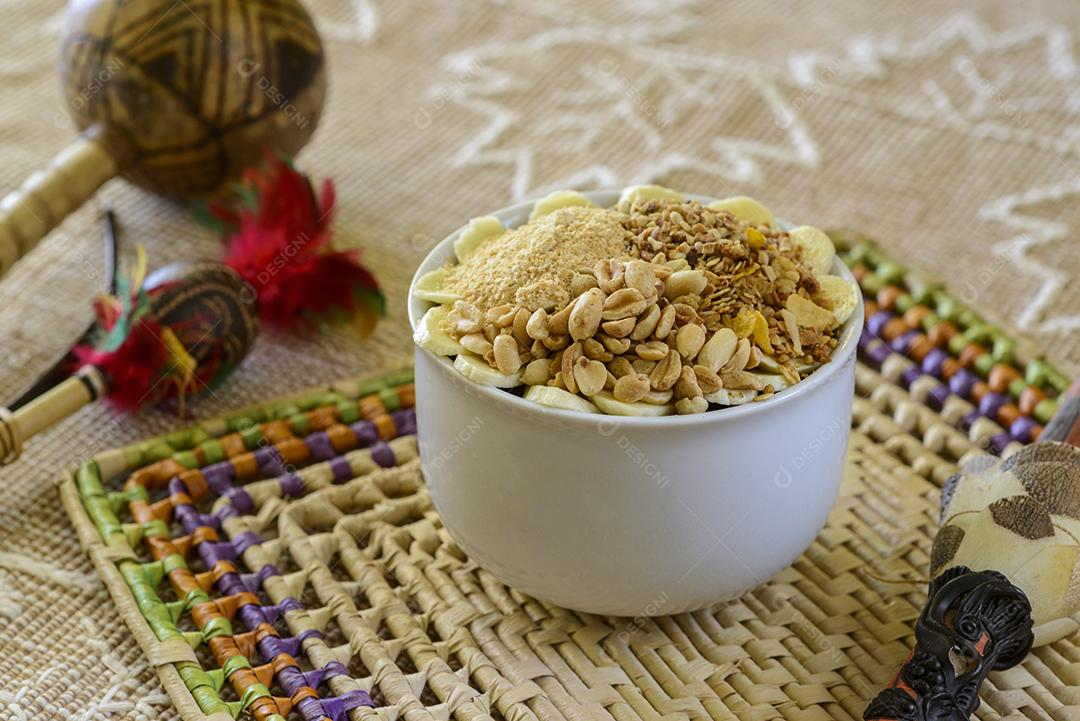 Tigela de açaí brasileiro com granola, amendoim e banana em uma mesa com artesanato da região amazônica borrada ao fundo.