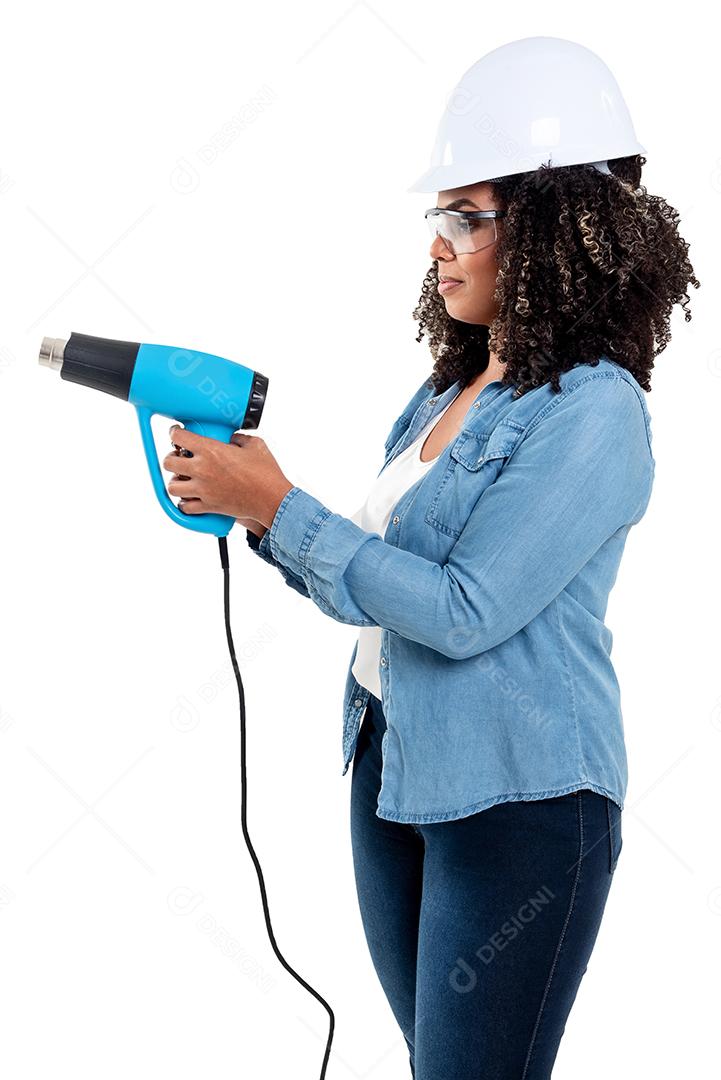 Mulher negra brasileira alegre e sorridente isolada de fundo branco, trabalhando com uma arma de calor usando óculos e um capacete na cabeça.