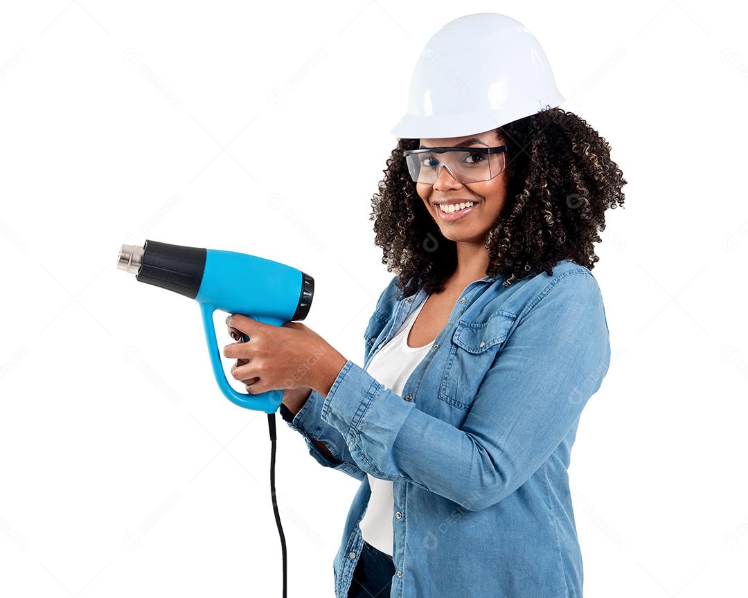 Mulher negra brasileira alegre e sorridente isolada de fundo branco, trabalhando com uma arma de calor usando óculos e um capacete na cabeça.