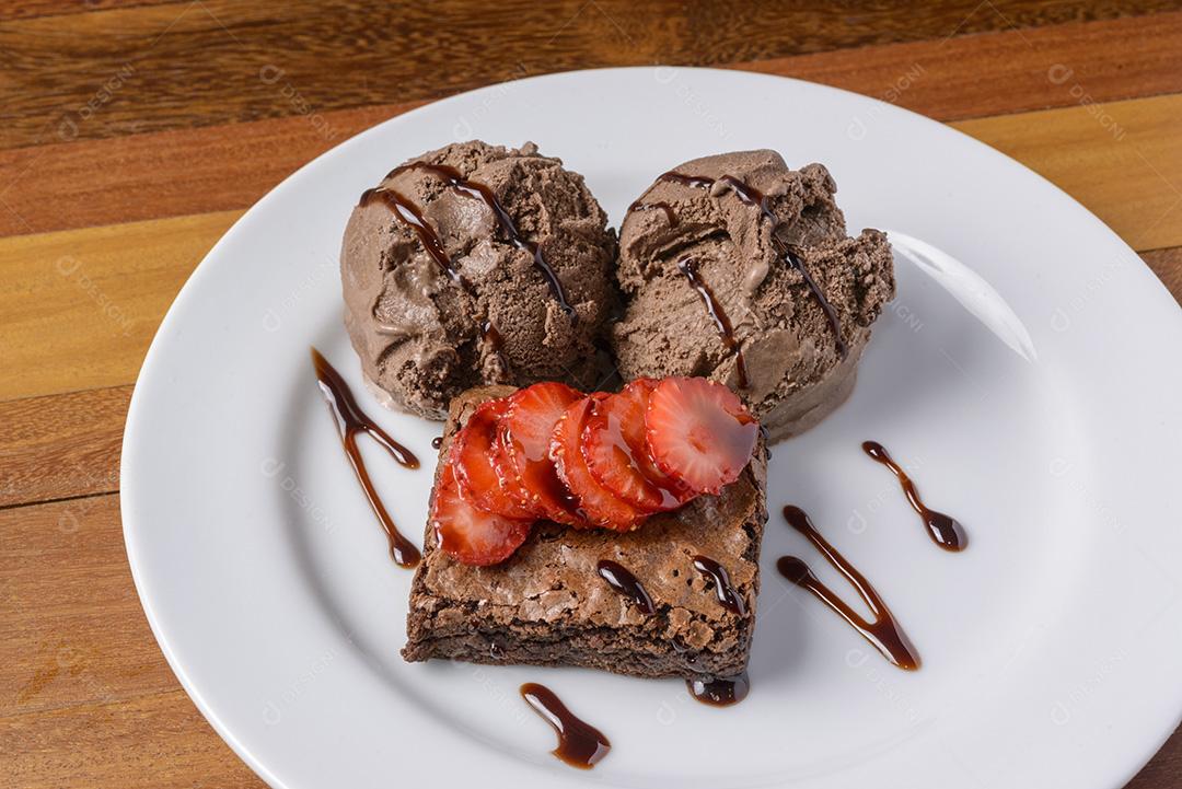 Brownie com morangos fatiados e sorvete de chocolate em um prato branco em uma mesa de madeira.