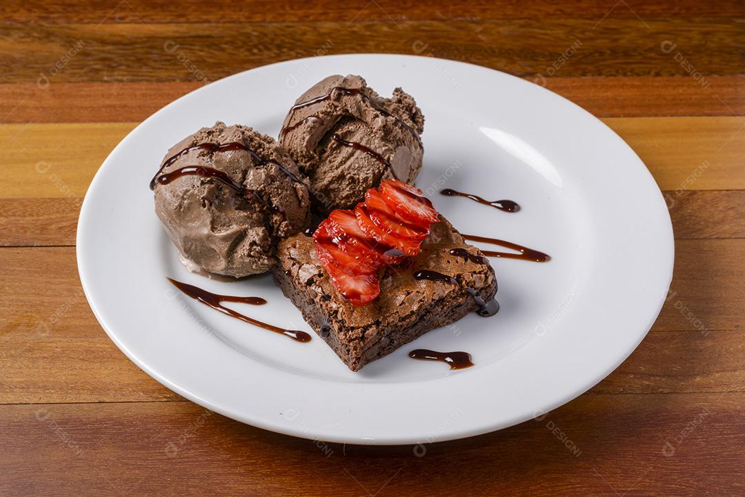 Brownie com morangos fatiados e sorvete de chocolate em um prato branco em uma mesa de madeira.