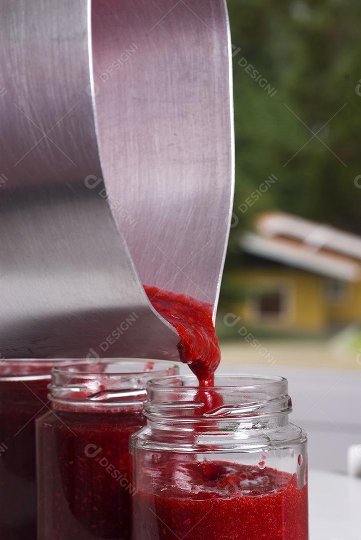 Panela botando geleia de morango dentro de uma frasco de vidro