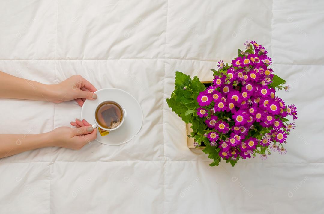 Braço de mulher com uma xícara de chá preto e flores roxas na cama branca.