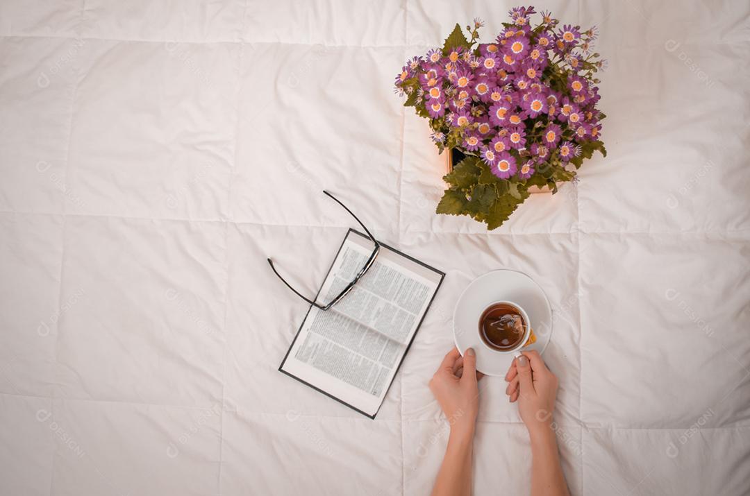 Braço de mulher com uma xícara de chá preto, óculos, livro e flores roxas na cama branca.