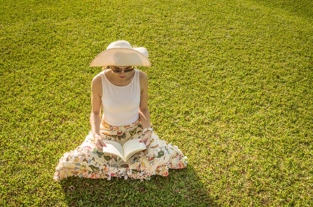 Linda mulher lendo livro sentado no gramado, visto de cima com chapéu de verão.