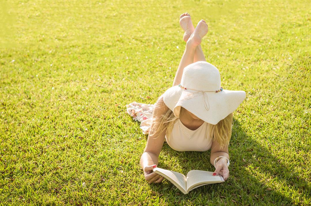 Linda mulher lendo livro deitado no gramado, visto de cima com chapéu de verão.