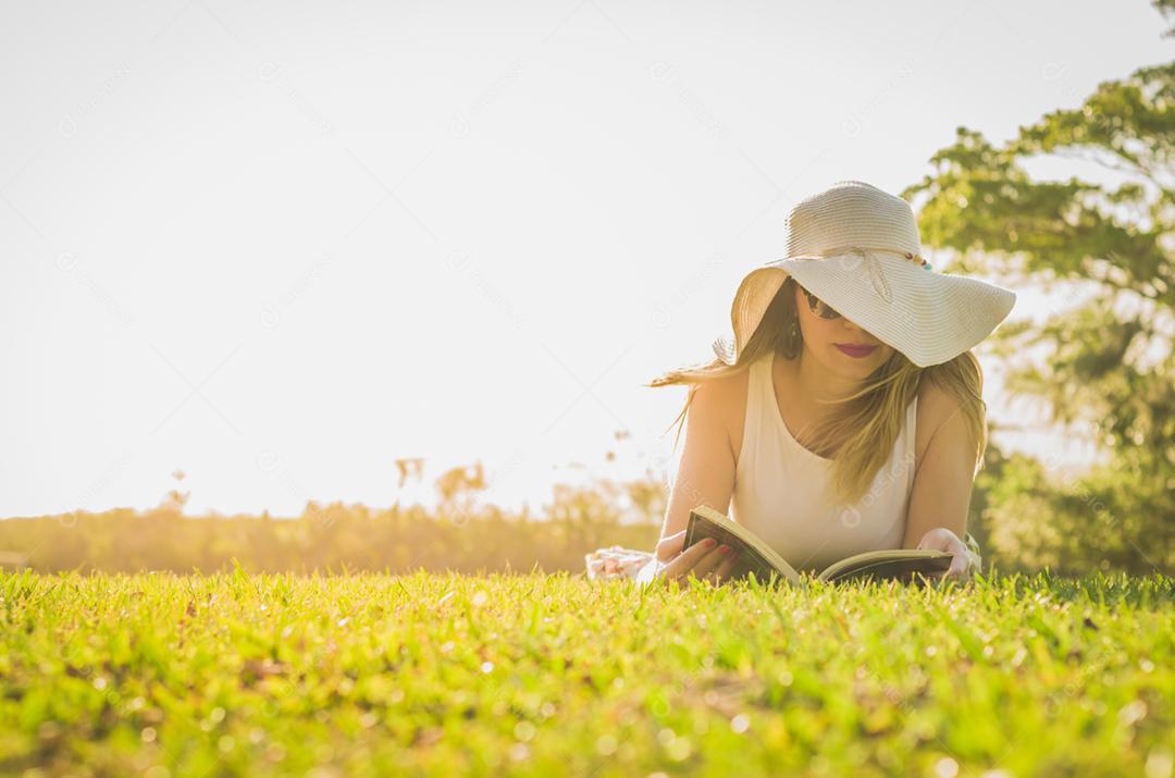 Linda mulher lendo livro deitado no gramado, visto de cima com chapéu de verão.