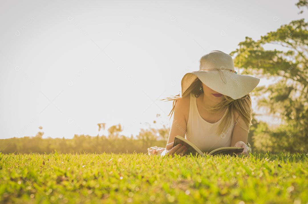 Linda mulher lendo livro deitado no gramado, visto de cima com chapéu de verão.