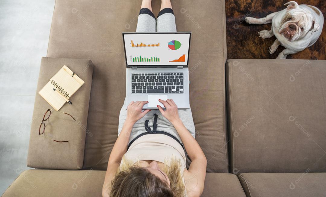 Mulher trabalhando em casa no computador, no sofá, animal de estimação sentado ao lado, bulldog inglês.