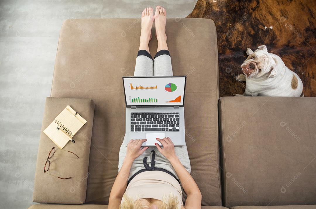 Mulher trabalhando em casa no computador, no sofá, animal de estimação sentado ao lado, bulldog inglês.