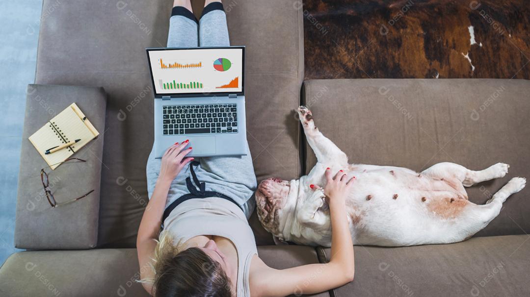 Mulher trabalhando em casa no computador, no sofá, animal de estimação sentado ao lado, bulldog inglês.