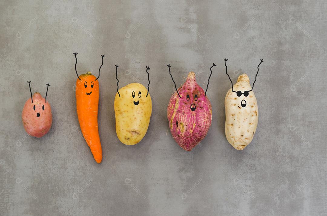 Legumes com rostos felizes com os braços para cima, batata, batata doce, cenoura. Fundo cinza, concreto polido.