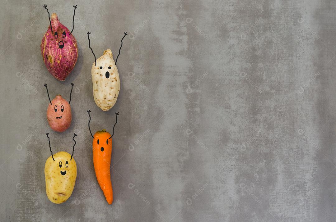 Legumes com rostos felizes com os braços para cima, batata, batata doce, cenoura. Fundo cinza, concreto polido.