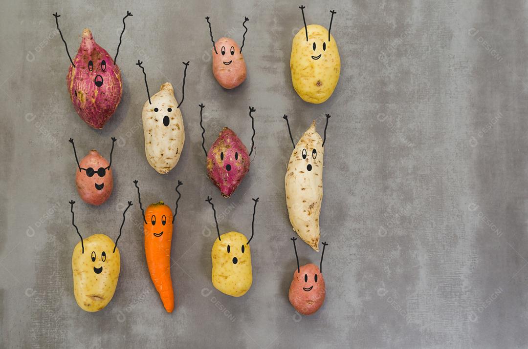 Legumes com rostos felizes com os braços para cima, batata, batata doce, cenoura. Fundo cinza, concreto polido.