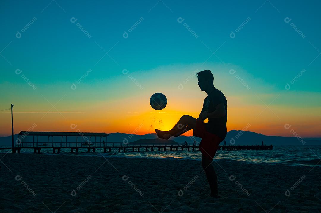 Homem jogando bola em praia com por do sol lindo. Esporte. Futebol