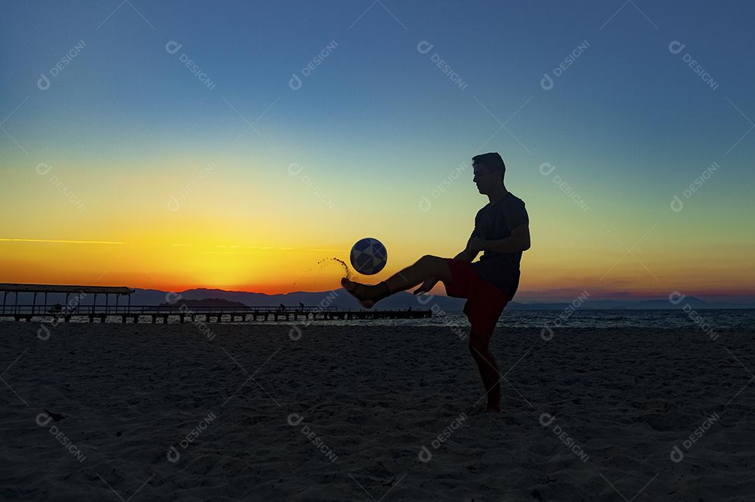 Homem jogando bola em praia com por do sol lindo. Esporte. Futebol