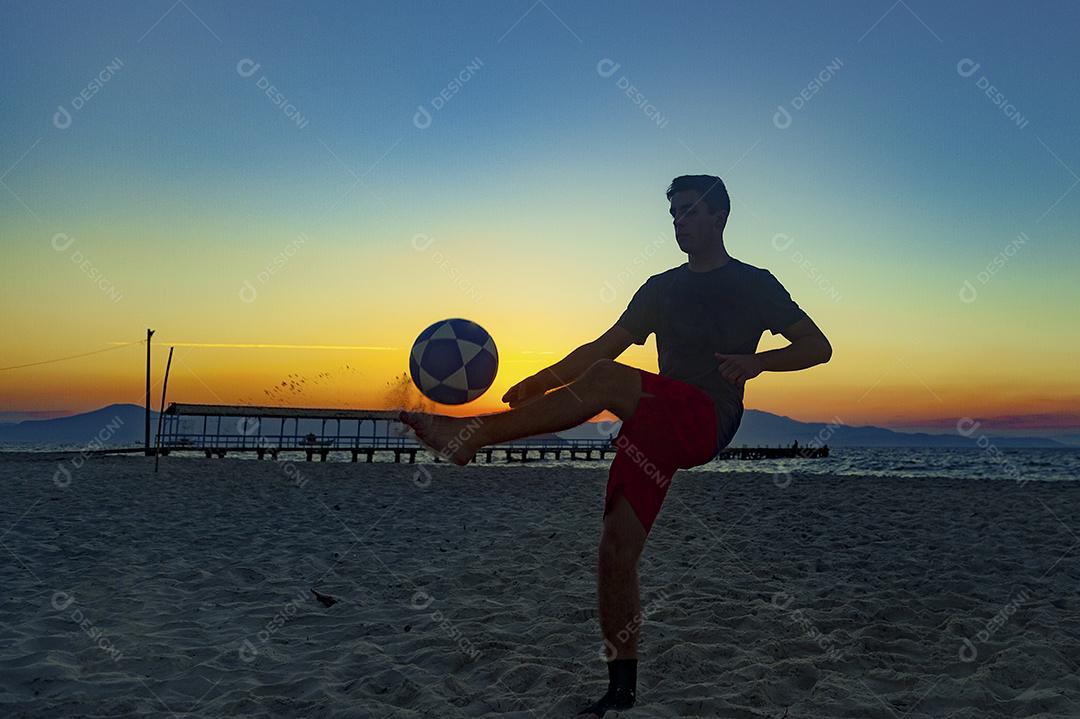 Homem jogando bola em praia com por do sol lindo. Esporte. Futebol