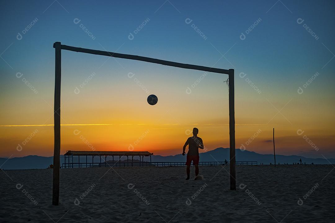 Homem jogando bola em praia com por do sol lindo. Esporte. Futebol