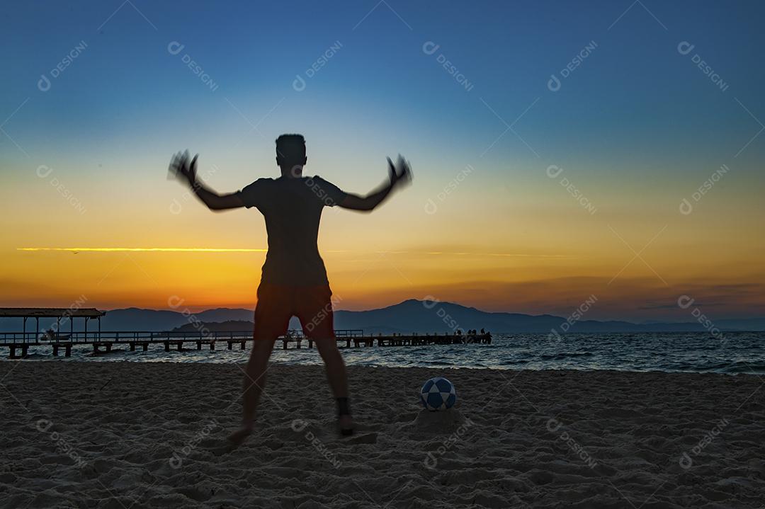 Homem jogando bola em praia com por do sol lindo. Esporte. Futebol