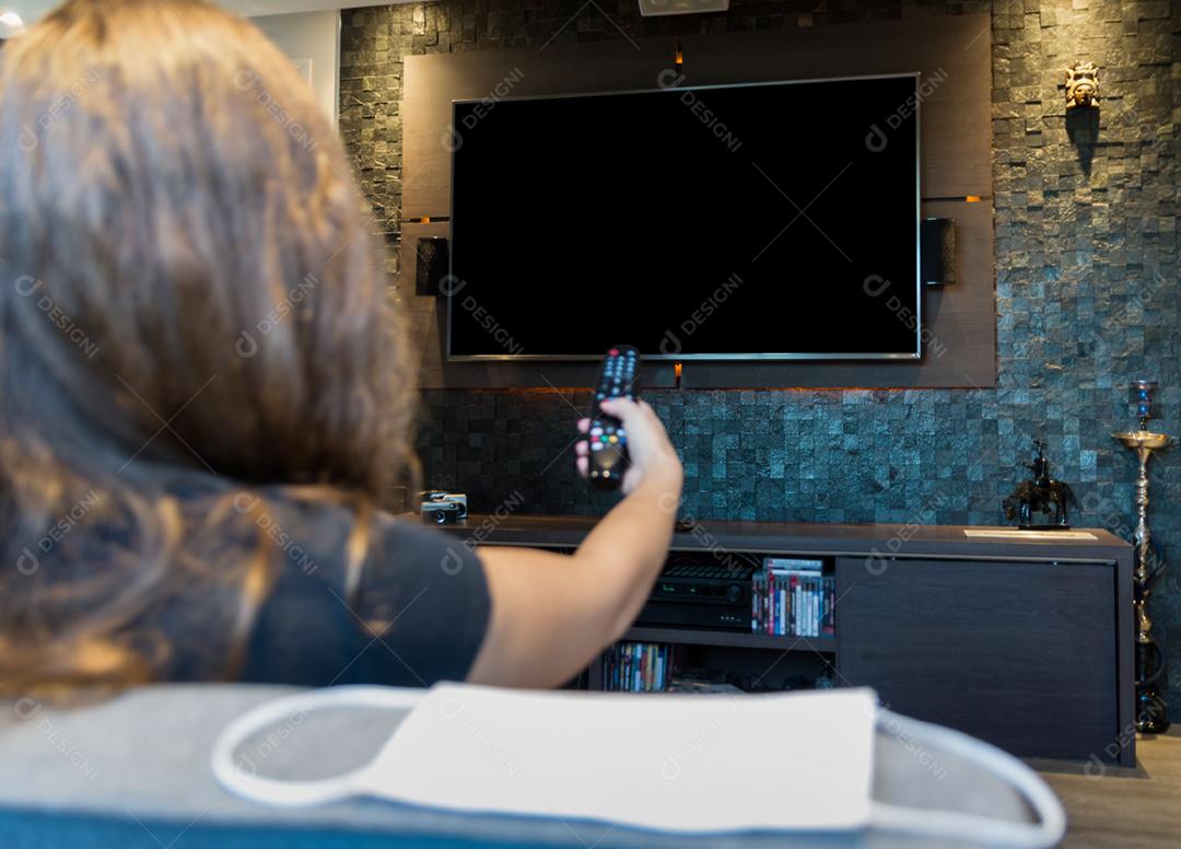 Jovem mulher trocando canais de TV com controle remoto, máscara protetora deixada no canto do sofá.