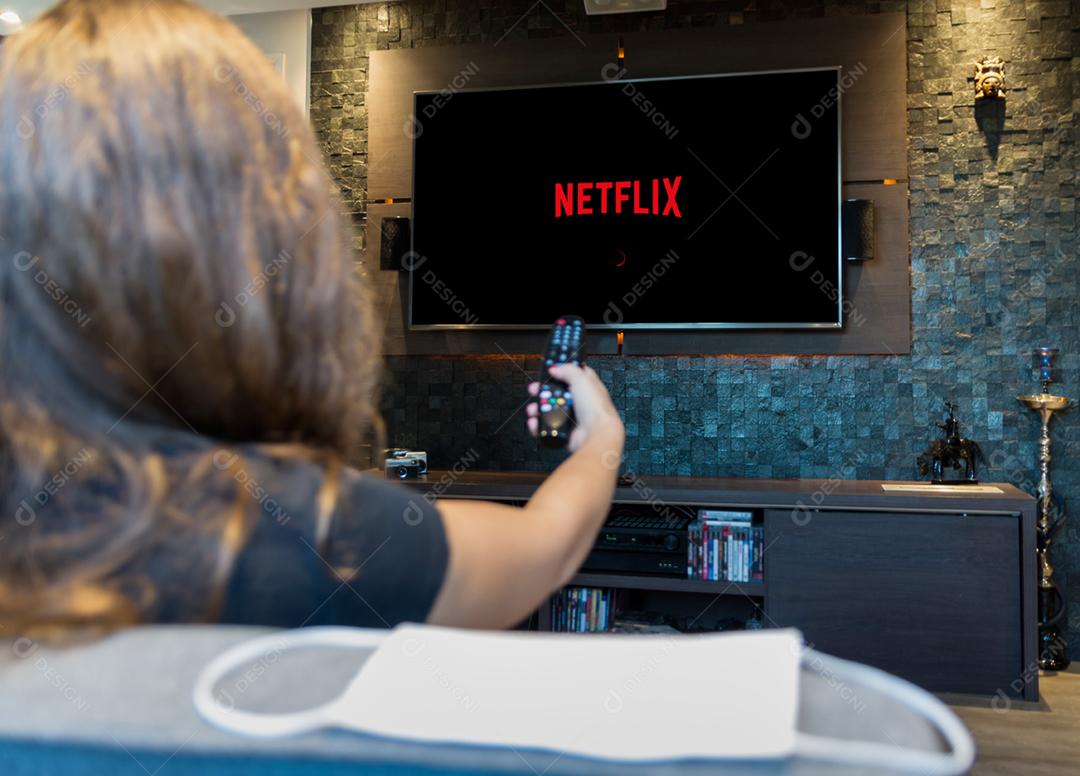 Jovem mulher trocando canais de TV com controle remoto, máscara protetora deixada no canto do sofá.