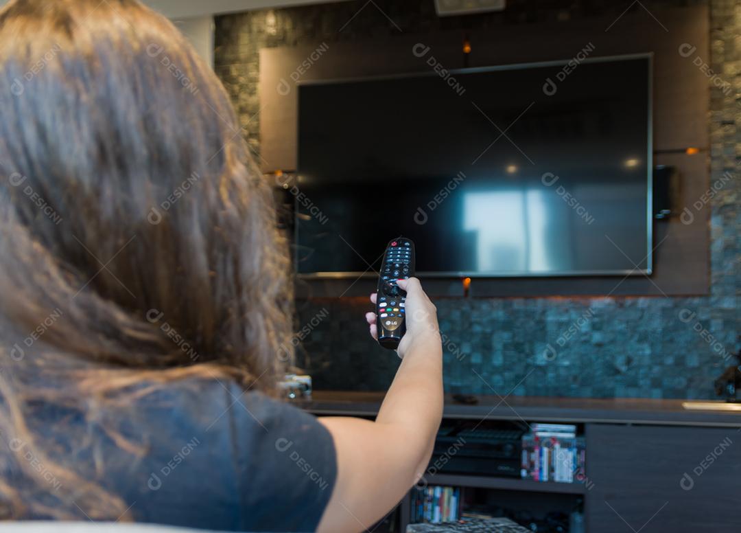 Jovem mulher trocando canais de TV com controle remoto, máscara protetora deixada no canto do sofá.