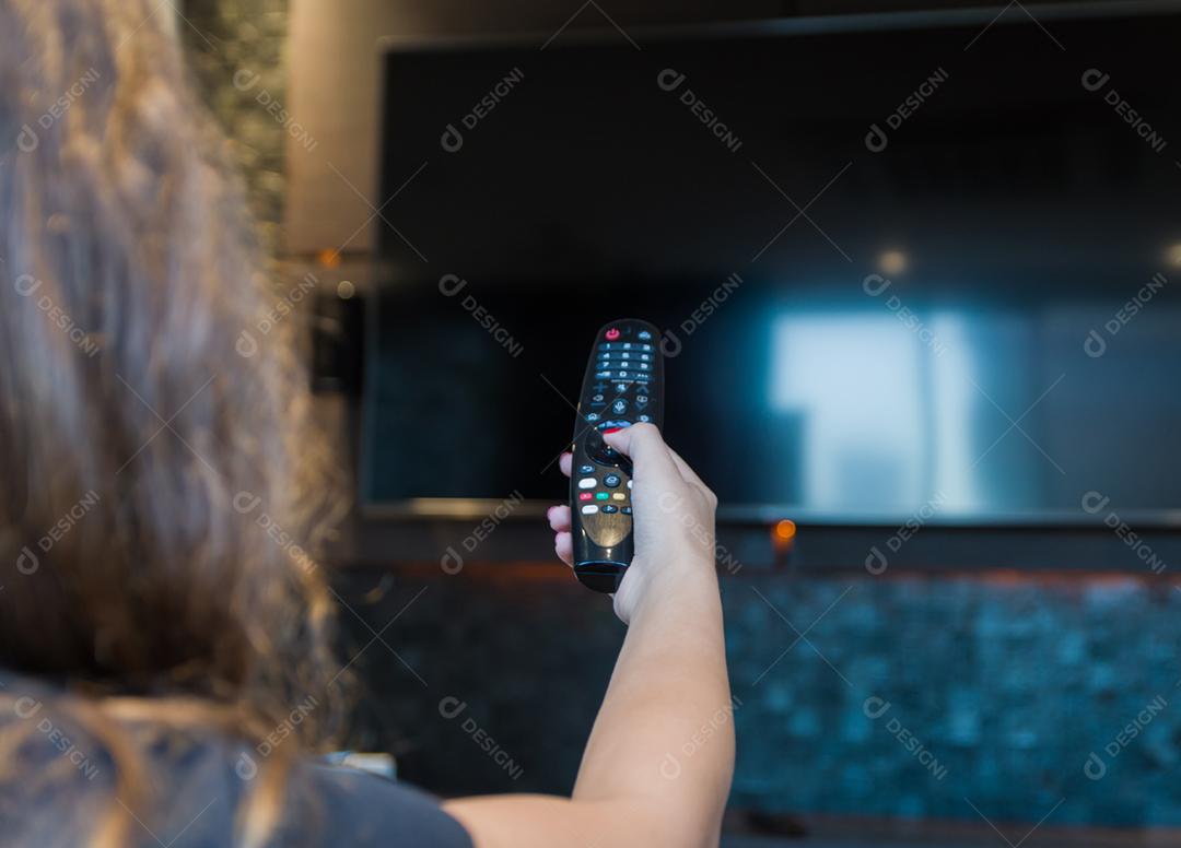 Jovem mulher trocando canais de TV com controle remoto, máscara protetora deixada no canto do sofá.