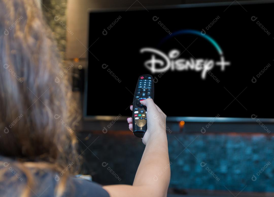 Jovem mulher trocando canais de TV com controle remoto, máscara protetora deixada no canto do sofá.