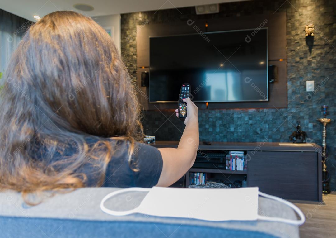 Jovem mulher trocando canais de TV com controle remoto, máscara protetora deixada no canto do sofá.