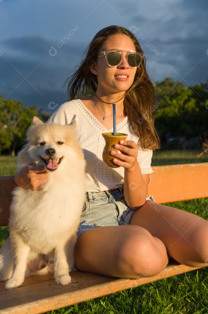 Mulher loira tomando chimarrao tradicional gaúcho com cachorro no parque.