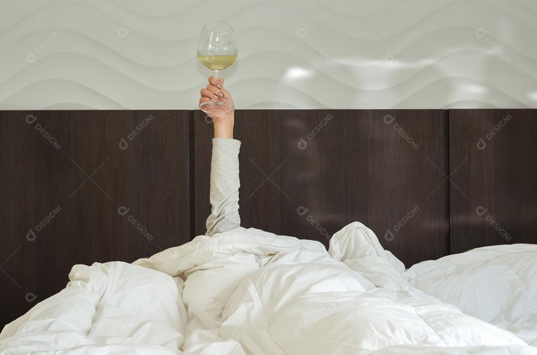 Mulher na cama, debaixo do edredom, com o braço levantado segurando o copo de vinho